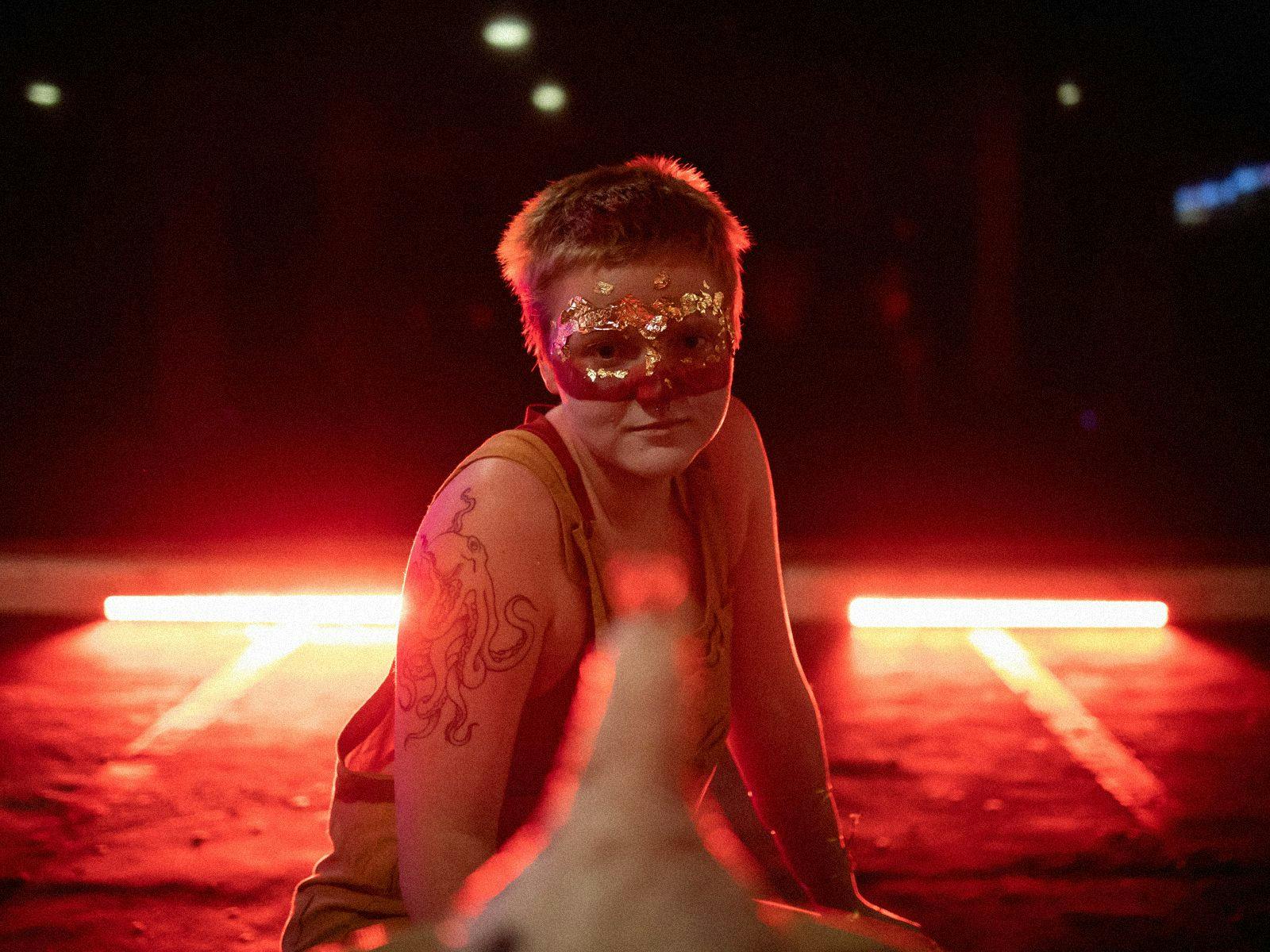 A figure sits crouched on concrete wearing a sparkly mask, red lights light up the darkness behind them.