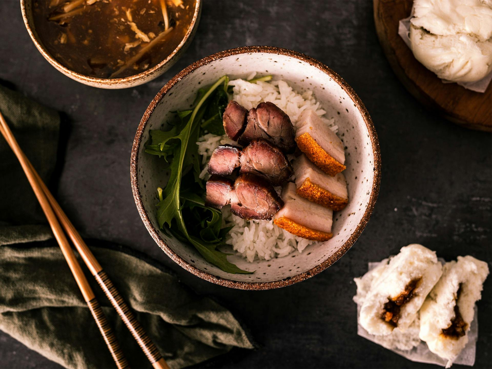 A feast of pork belly and char siu, pork buns and hot and sour soup on a table with chopsticks and a linen napkin