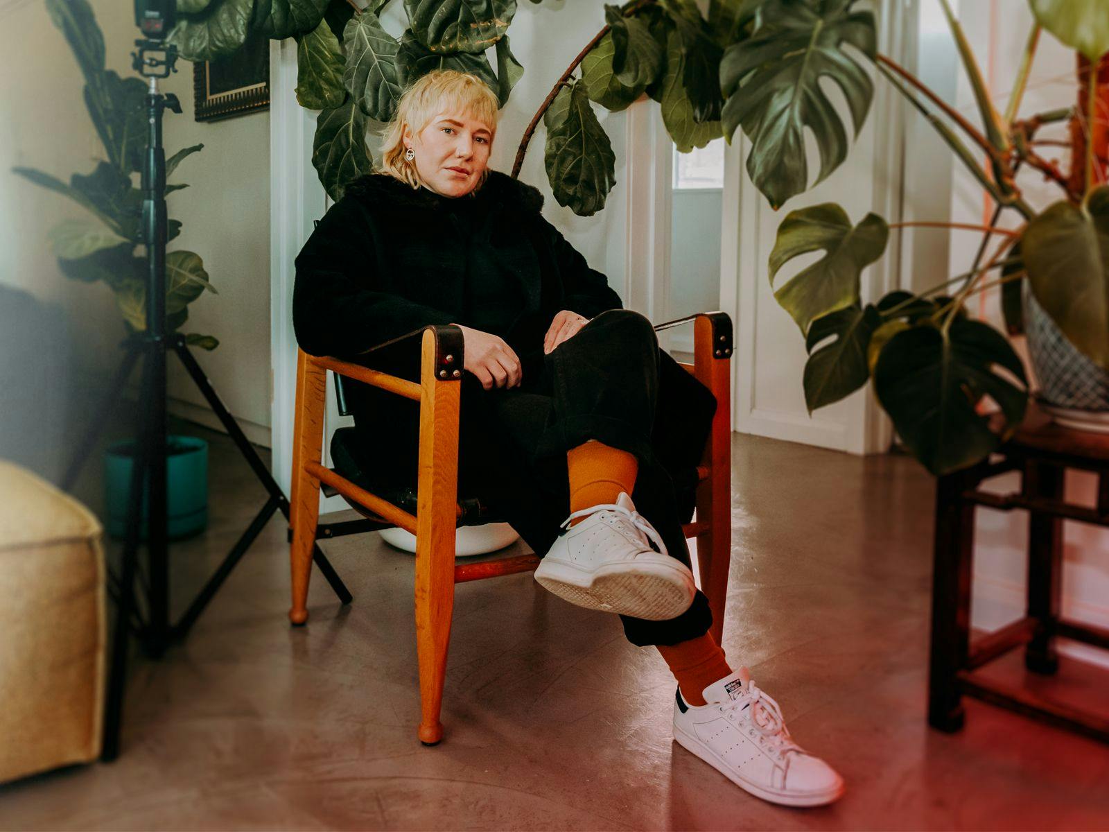 ONYX sits on a wooden chair, several large leafy indoor plants surround them.