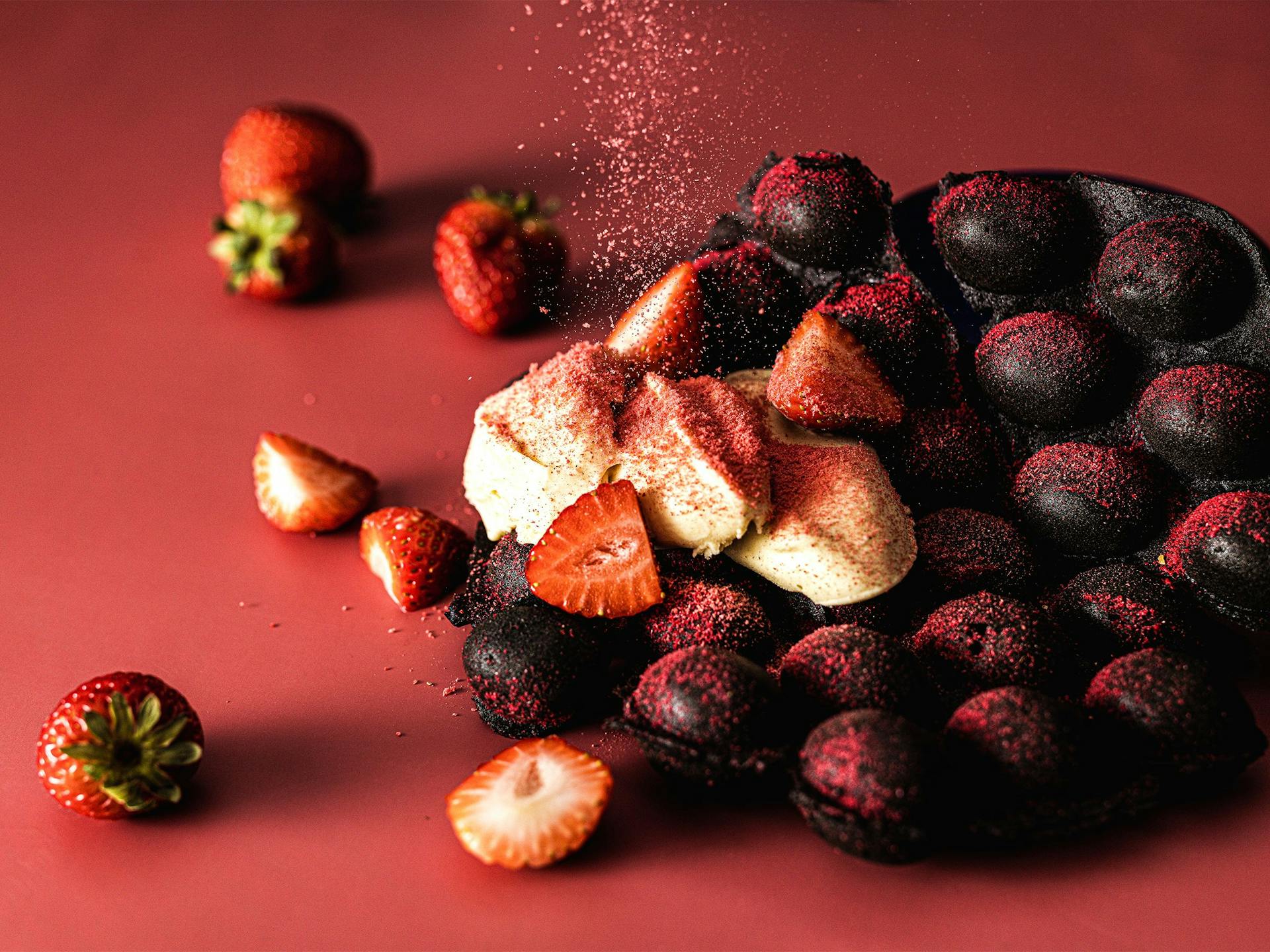 A chocolate eggpuff waffle with ice cream and strawberries, being sprinkled with freeze dried strawberry powder