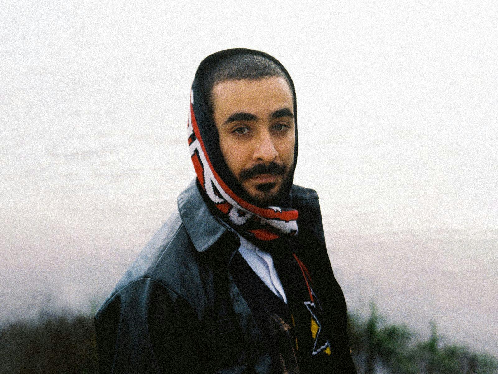 Moktar wearing a leather jacket, with a red and white head scarf.