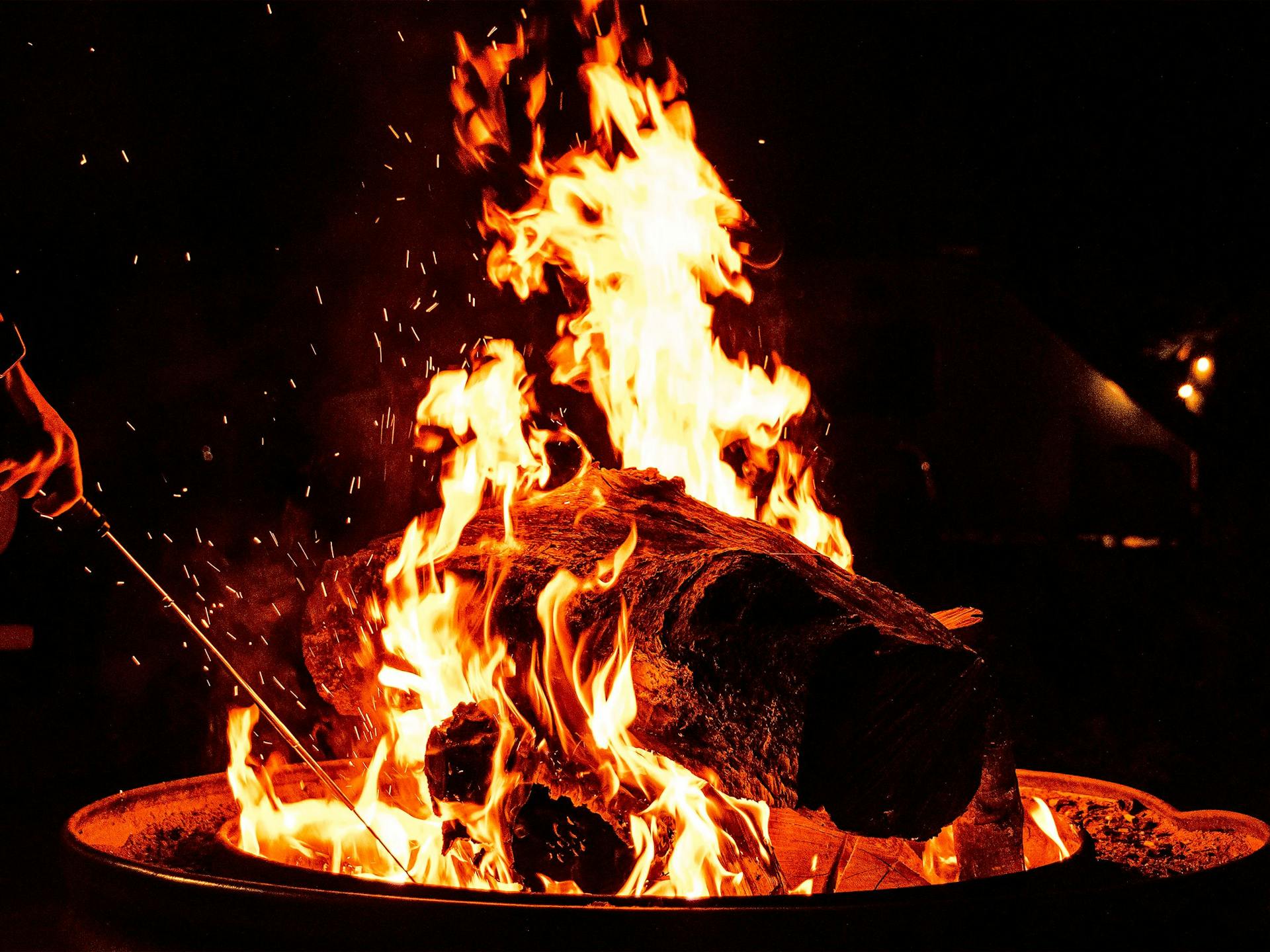 A raging fire in a fire pit with a big log and someone poking it with a stick