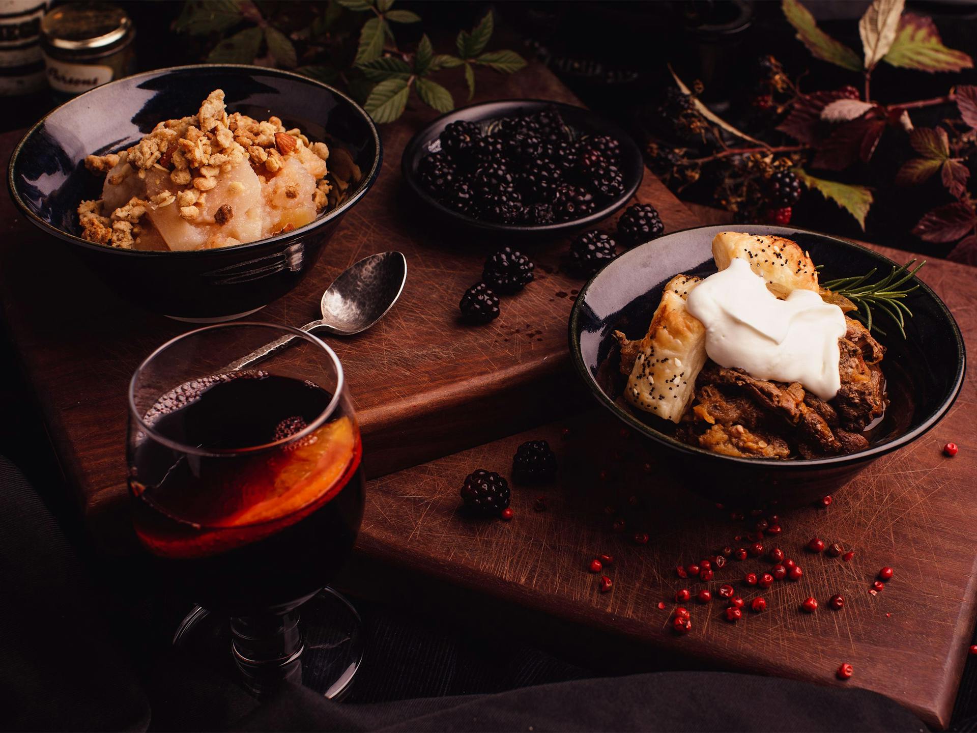 A feast of desserts and berries sit on a wooden board next to mulled wine and leaves