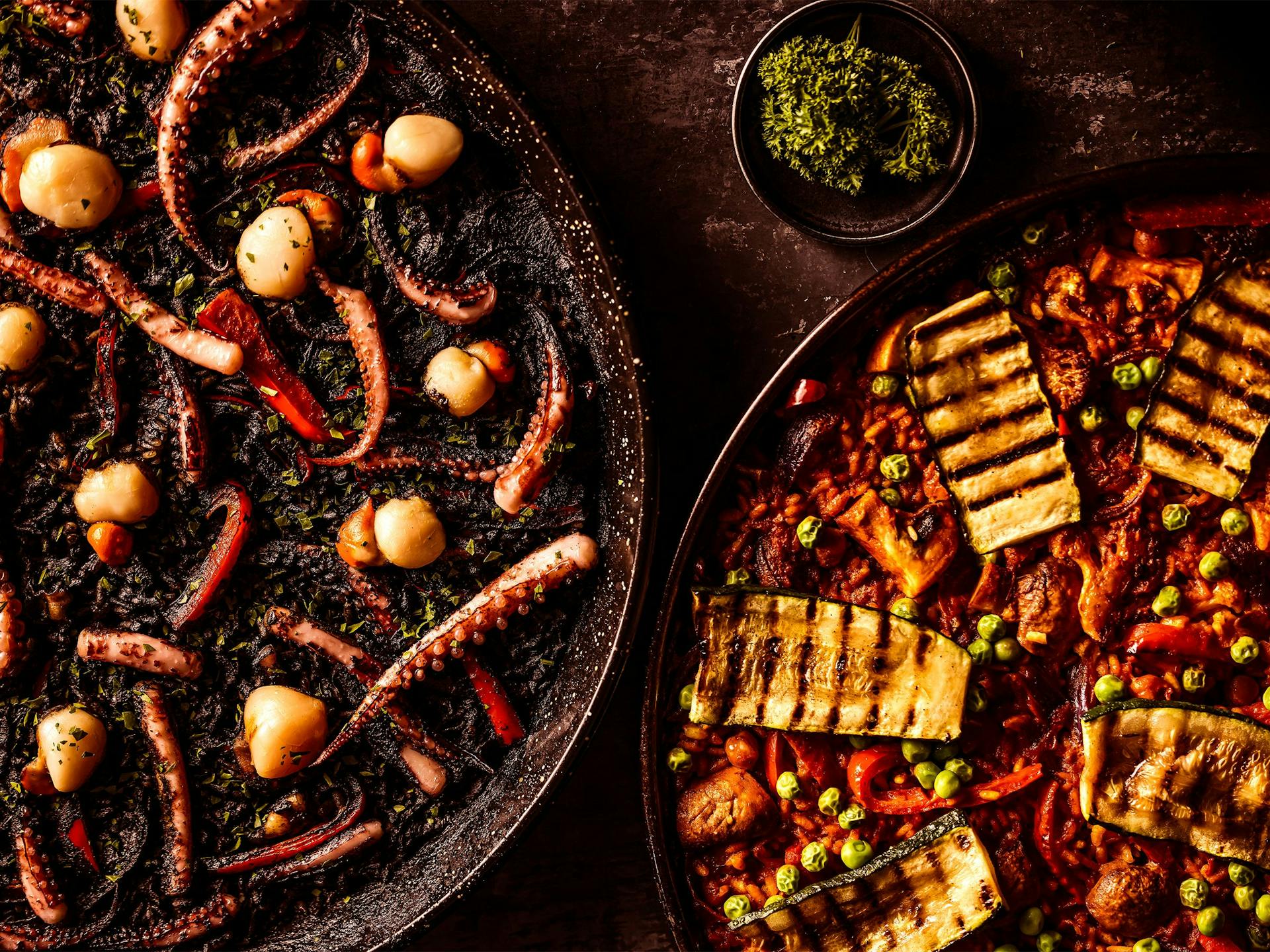 A black seafood paella and a vegetable paella sit asymmetrically in the image in large dishes, with a small bowl of parsley next to them