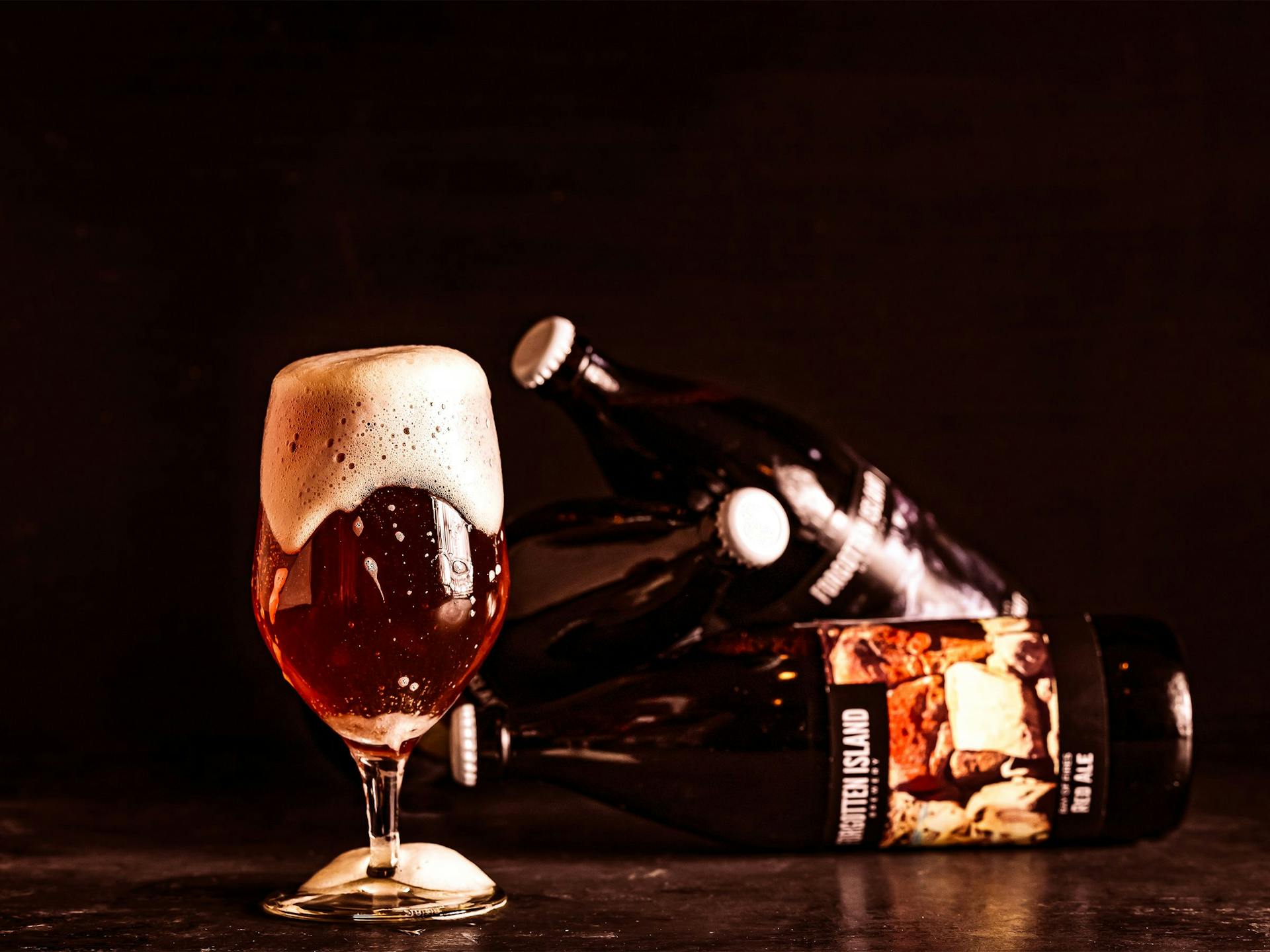 An overflowing glass of beer sitting next to three beer bottles balancing in the background