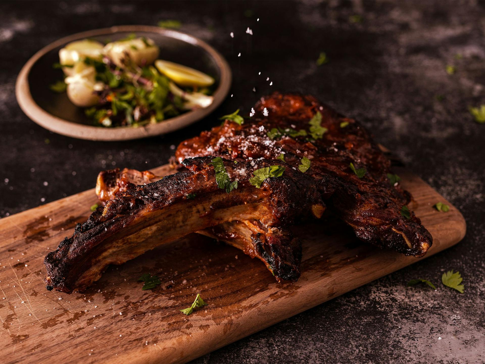 Salt is being sprinkled on beef short ribs that sit on a wooden board with potatoes and salad in the background