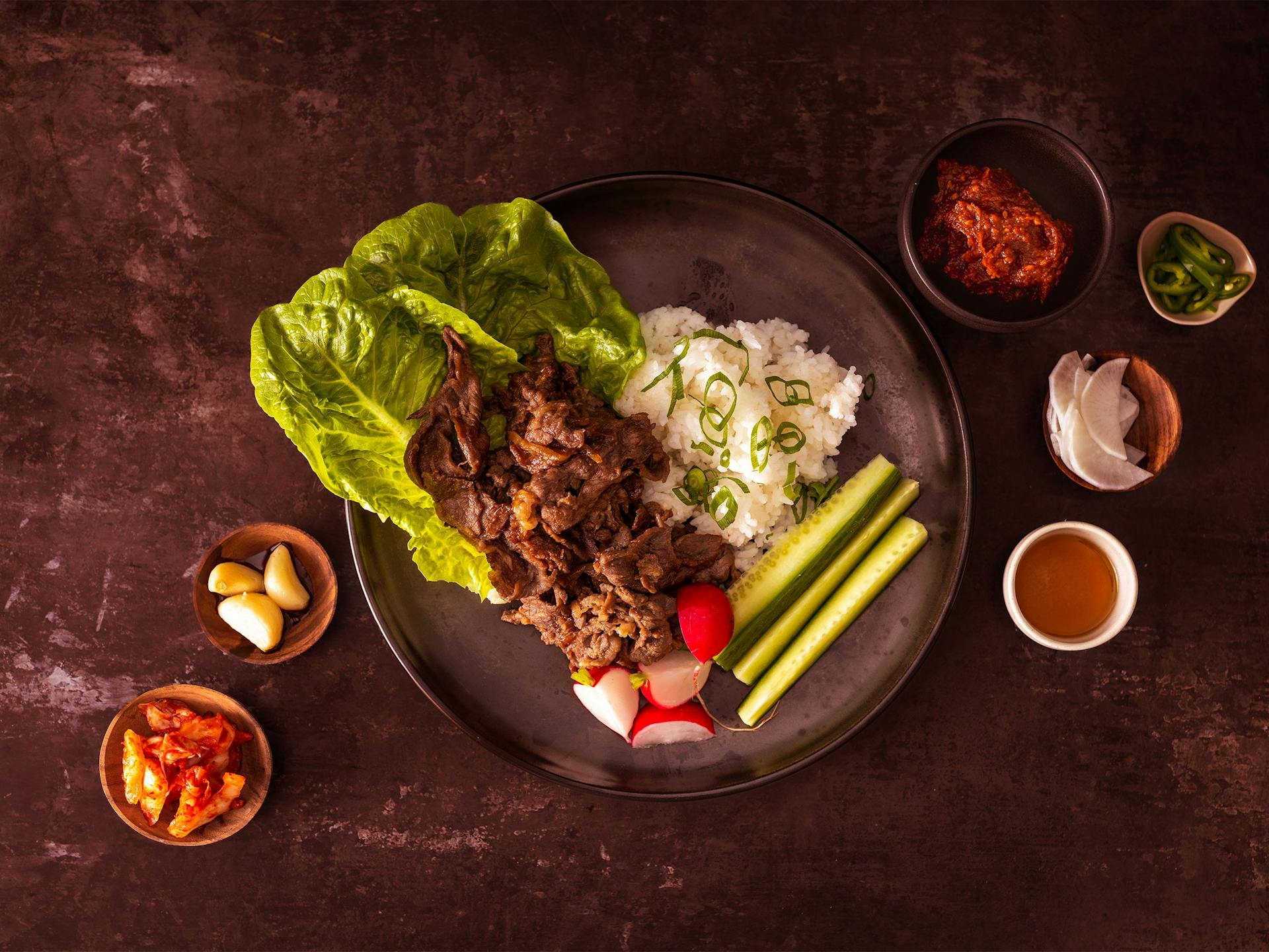 A bulgogi beef ssam plate surrounded by small side bowls of garlic, kimchi and more