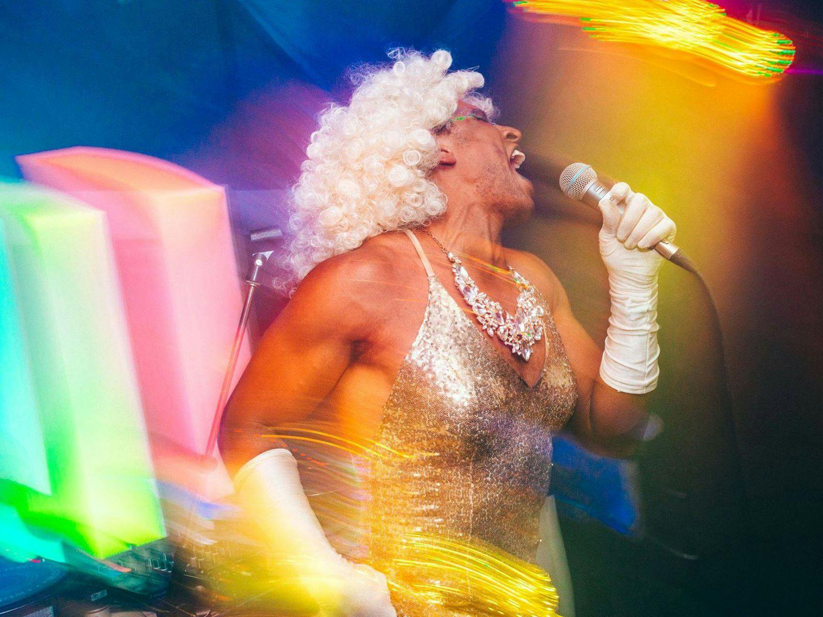 Nefertiti LaNegra wearing a silver dress and big white wig, singing into a microphone.