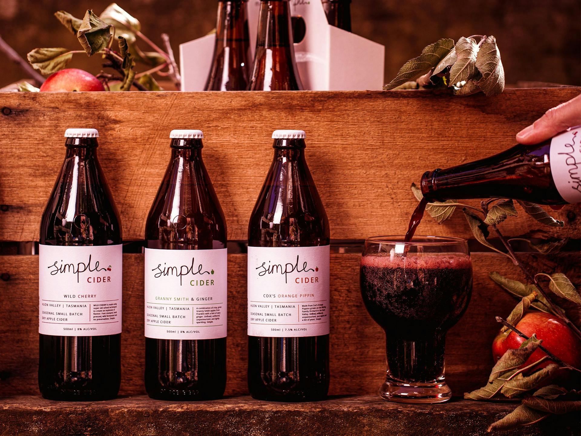 A glass and three bottles of cider sit against an apple crate