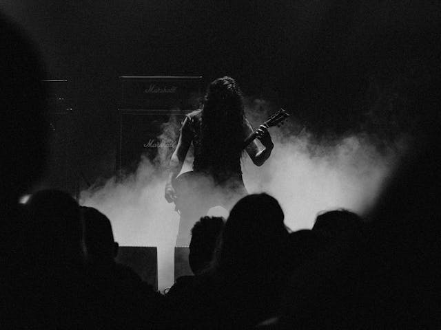 A mysterious guitar player stands alone on-stage, there is smoke and large speakers behind them.