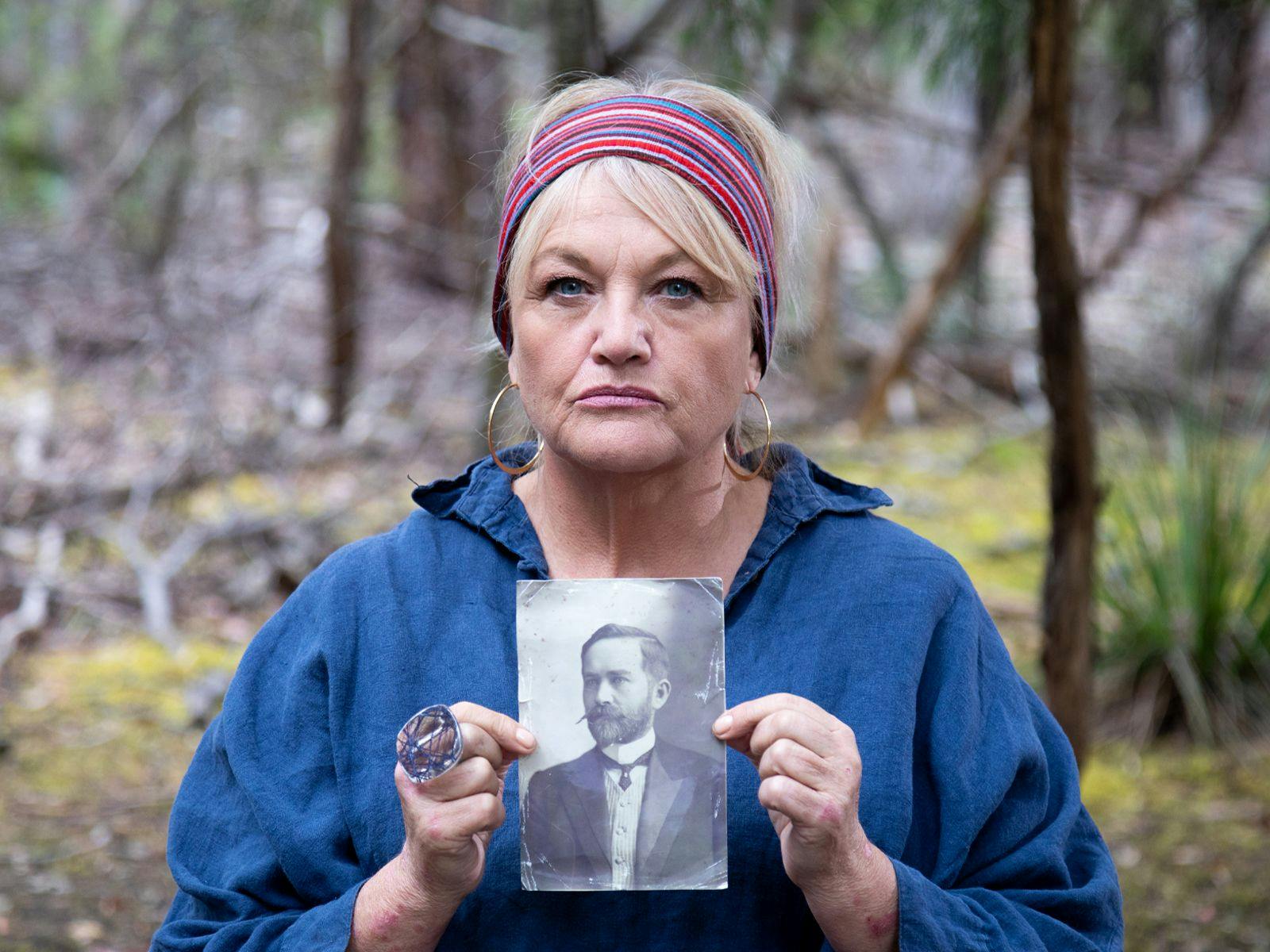 A person wearing a blue jacket, hoop earrings and striped headwear holds an old black and white printed photograph.