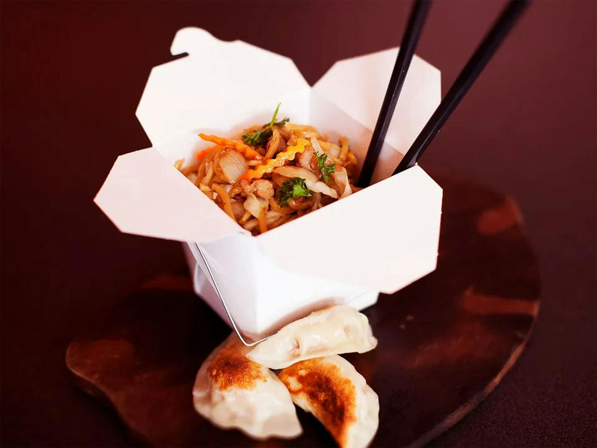 Stir-fried noodles in a takeaway box with chopsticks and some dumplings on the side