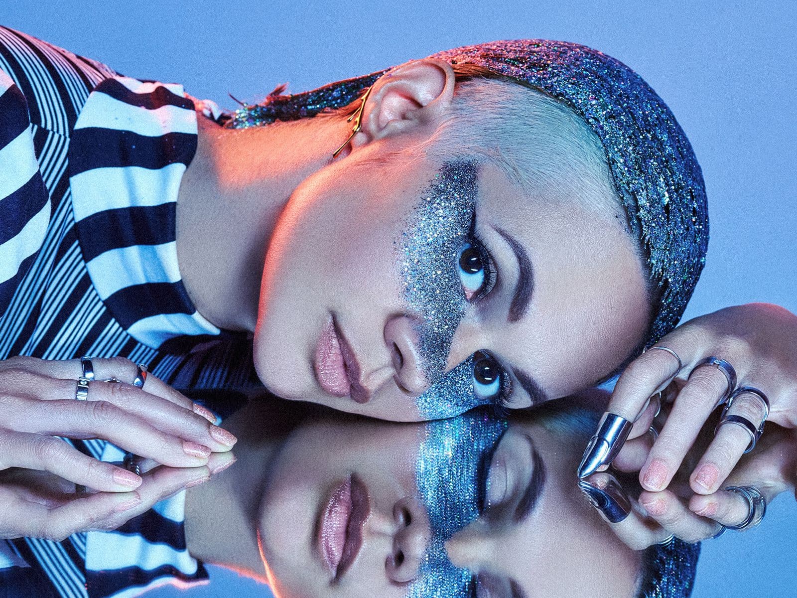 GLVES lying on their side on a mirrored surface. Blue sparkly hair and makeup dominate their appearance.