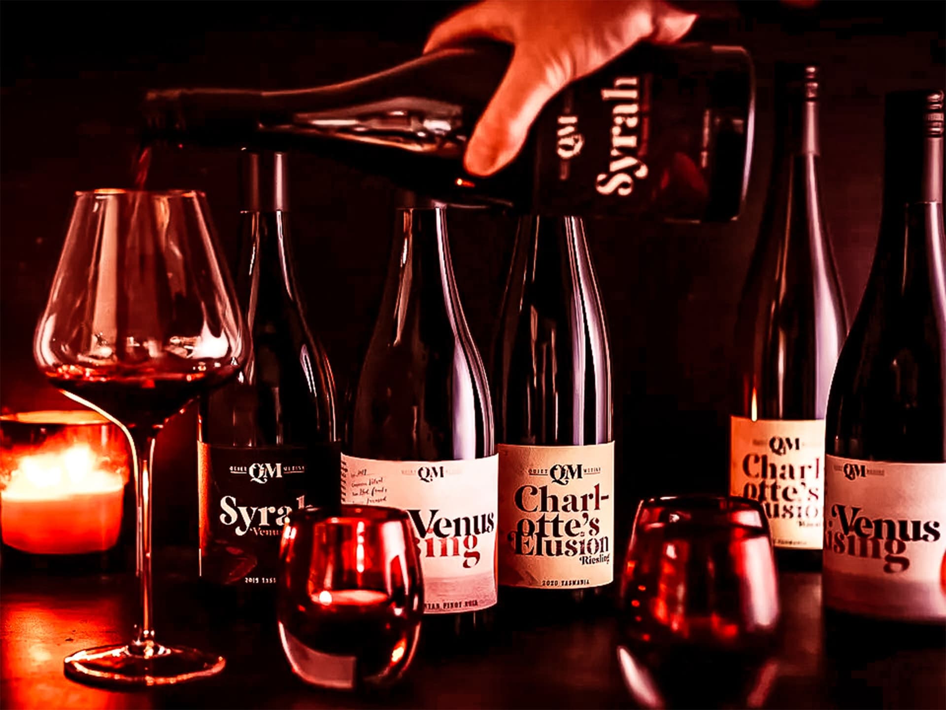 A hand pouring a glass of syrah next to five bottles of wine and candles