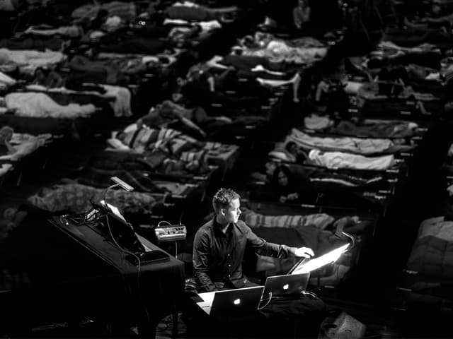Max Richter performing, with several people sleeping in beds behind him.